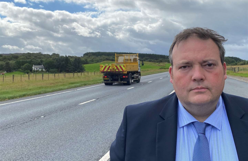 Jamie, wearing a suit, is standing beside the A9. A lorry is driving past.